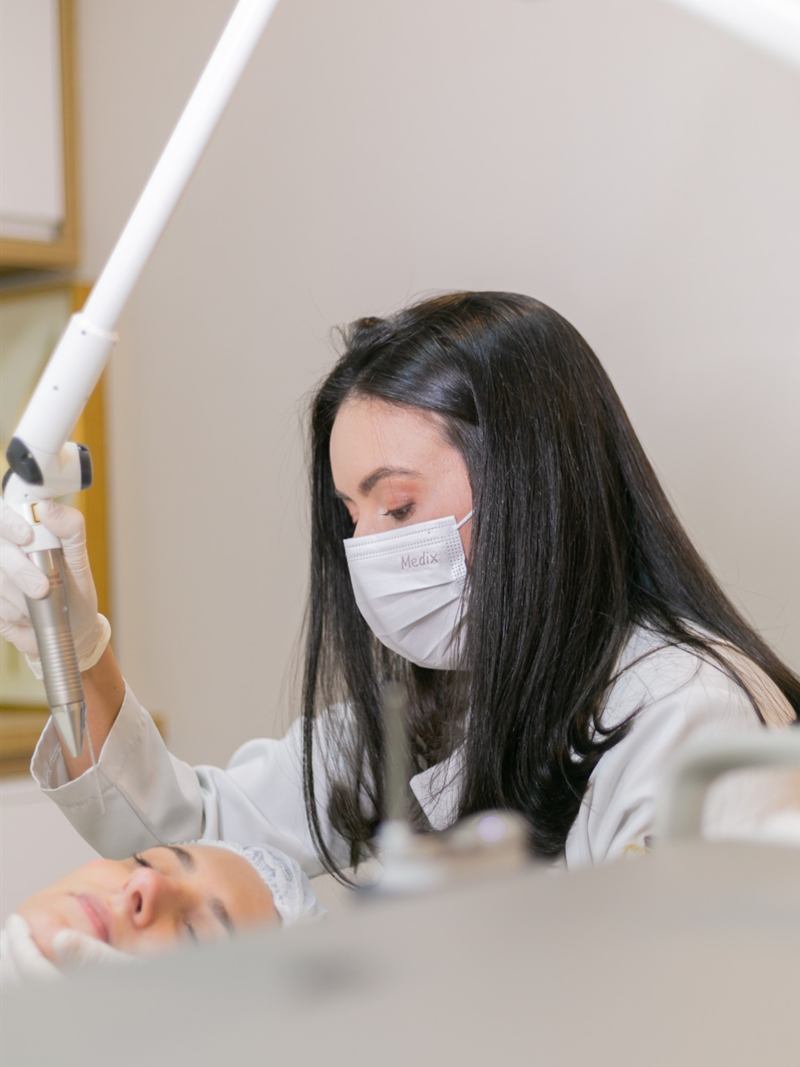 Médica de jaleco e máscara aplicando o laser Fotona no rosto de uma paciente, em consultório