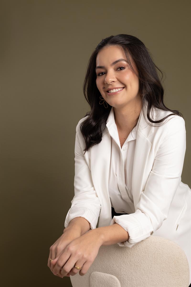 Dra. Thaís Gama sorrindo, de blazer branco, sentada e apoiada no encosto de uma poltrona, em fundo verde-oliva